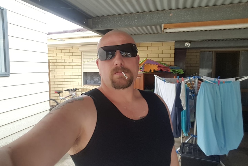 A man in a singlet and sunglasses takes a selfie.
