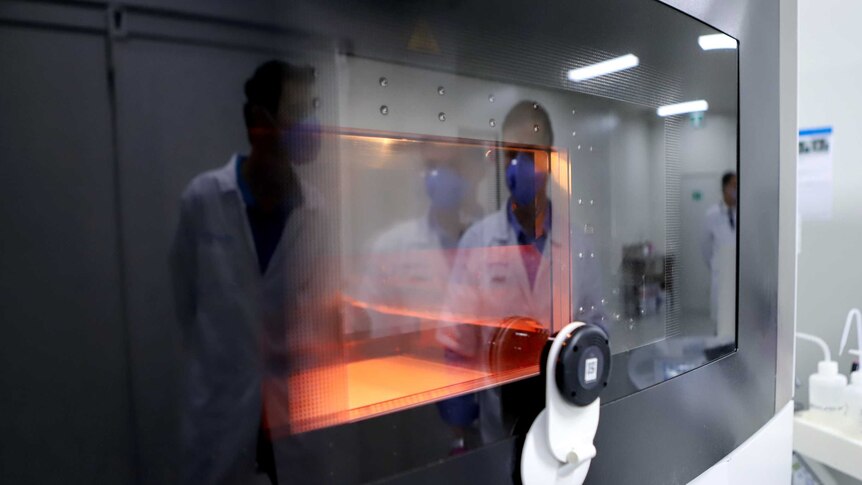 A glass screen with a orange furnace inside and a reflection of men in lab coats and mask in the glass