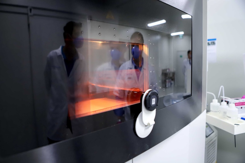 A glass screen with a orange furnace inside and a reflection of men in lab coats and mask in the glass