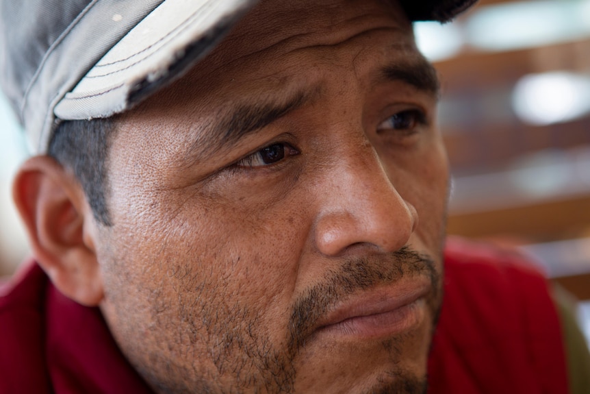 A close profile shot of a man with dark features and a light mostache wearing a cap and red top 