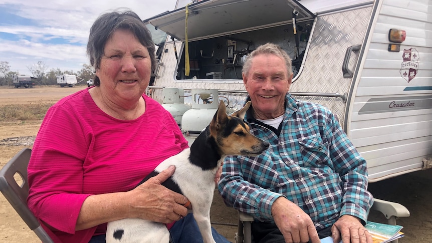 A couple sitting in front of their caravan, with their dog in their lap.