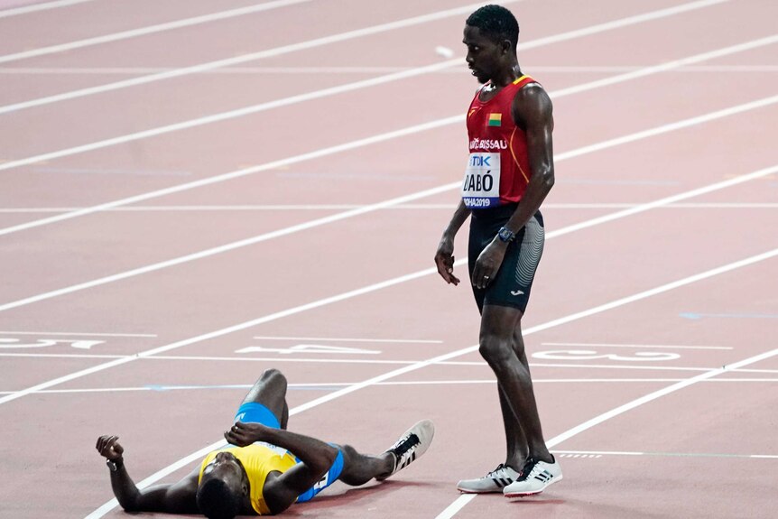 A male track and field athlete stands on the track next to another athlete who is lying on the track.