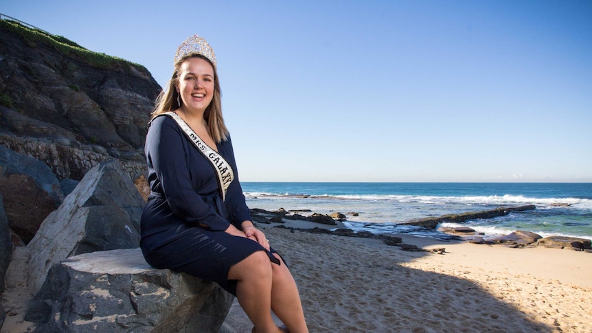 Emilie Hardes sits on a rock next to the beach wearing a crown and sash.