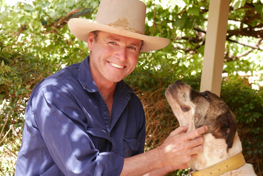 Brendan smiles at the camera and crouches next to a dog, patting it.