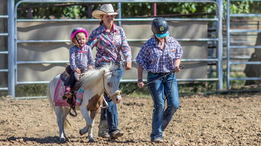 A little girl on her pony is led across what looks to be a rodeo arena.