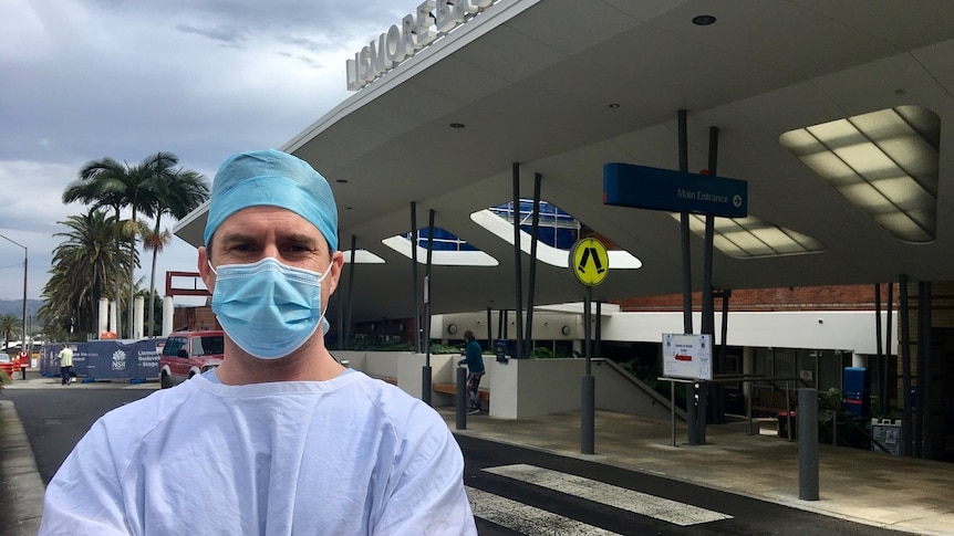 Male surgeon in pale blue scrubs, white apron, blue face mask and blue cap in front of hospital