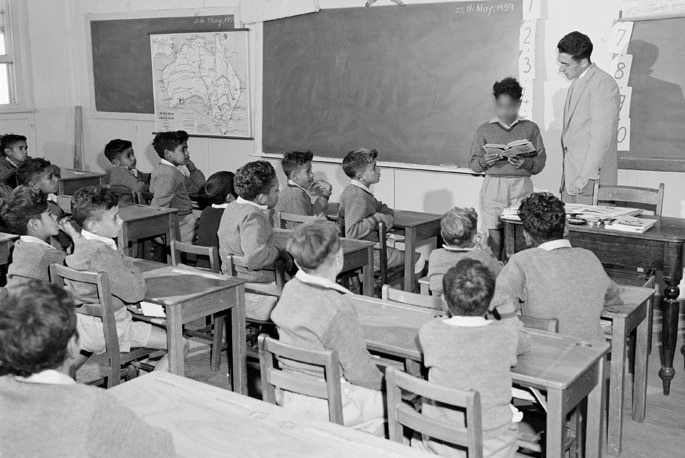 Young Aboriginal boys sit at desks in classroom, with a teacher standing before them.