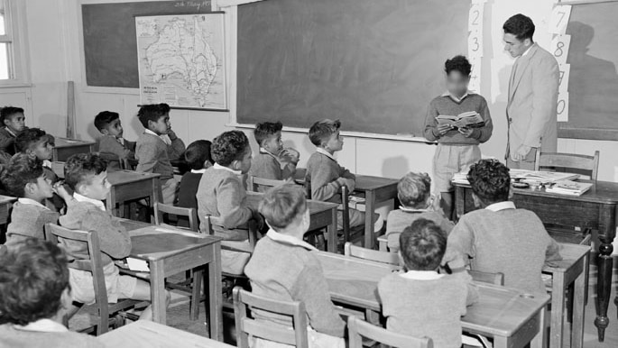 Young Aboriginal boys sit at desks in classroom, with a teacher standing before them.