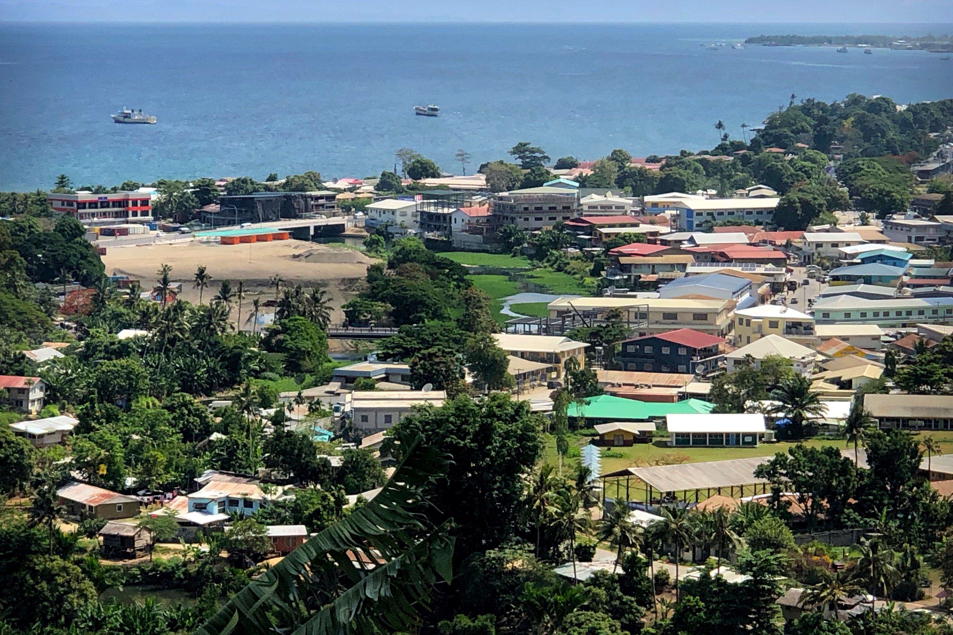 Ships are docked offshore from a small settlement on a verdant tropical island surrounded by light blue waters. 