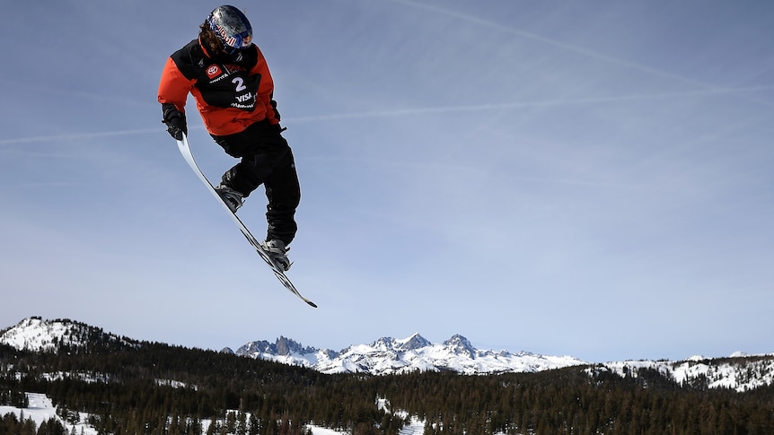 Val Guseli holds his board in the air with mountains and snow in the background