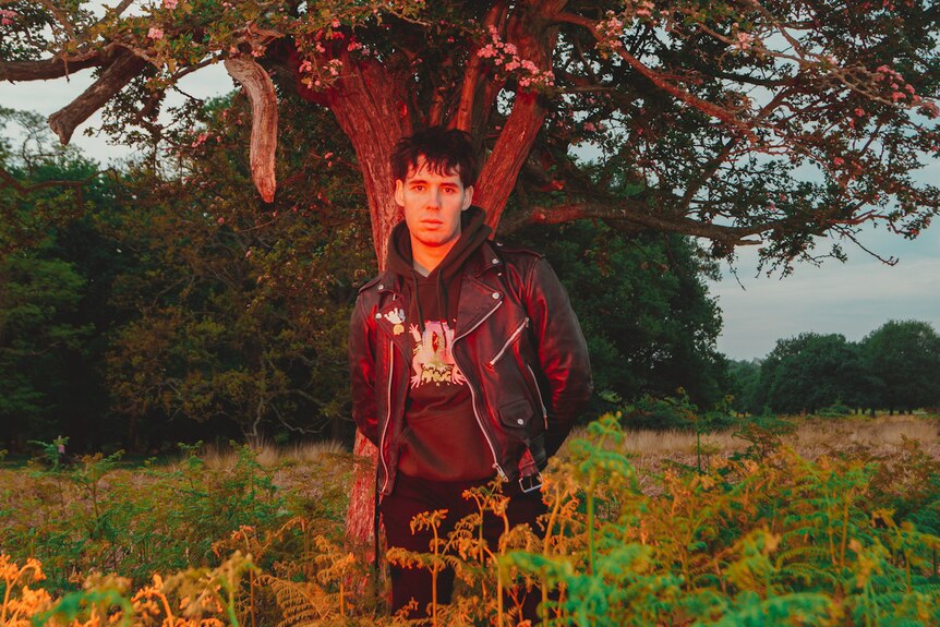 The artist wearing black leather jacket standing under tree in field of ferns at dusk, with red lighting from below.