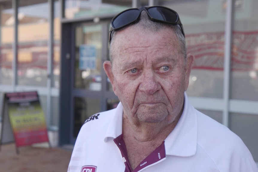 An elderly man in a white polo shirt and sunglasses looks away from camera, upset. 