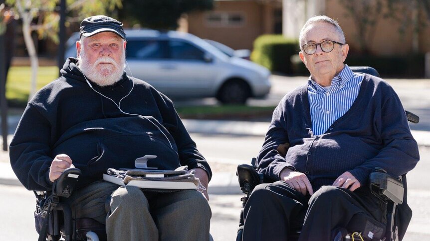 Two men in wheelchairs in front of a building with parked cars in the background.