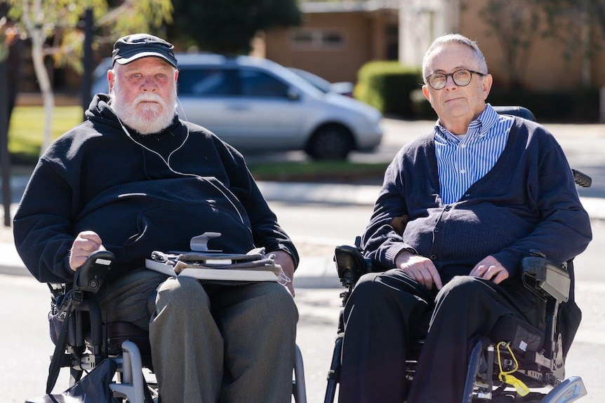 Two men in wheelchairs in front of a building with parked cars in the background.