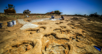 Wide divots in sandstone in the foreground, with people crouching in the dirt and bulldozers in the background under a blue sky.