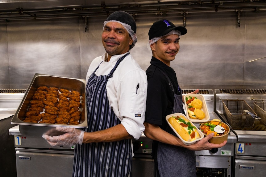 An older and younger man stand back to back, smiling, each holding trays of food.