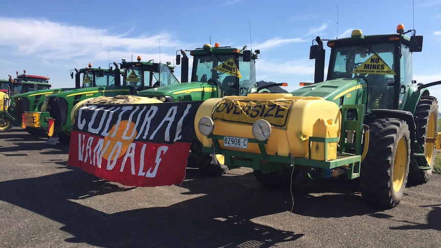 A line up of yellow and green tractors with one carrying an Indigenous flag with 'Cultural vandals' written on it.