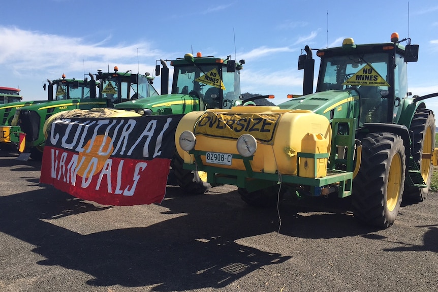 A line up of yellow and green tractors with one carrying an Indigenous flag with 'Cultural vandals' written on it.