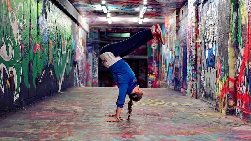 A female break-dancer doing a handstand move in the middle of a graffiti covered tunnel at night