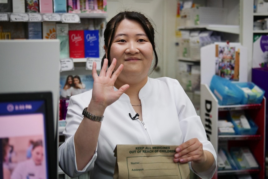 Judy Lam holds a brown paper bag, waving to a customer from behind the pharmacy counter.