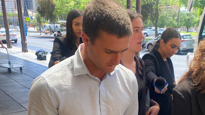 A man walks with his head down and hands in his pockets with a crowd of reporters around him