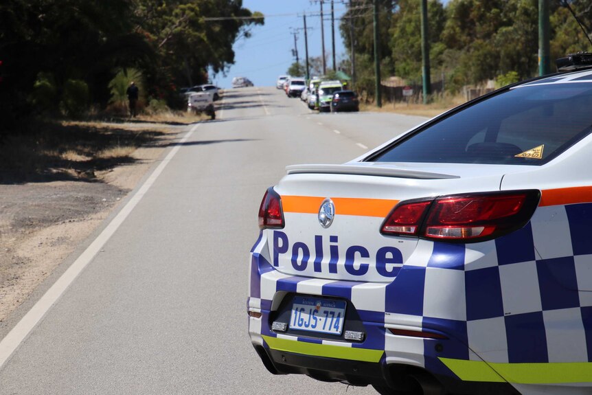 A WA Police car sits parked on a road lined by trees with other emergency vehicles on the side of the road in the distance.