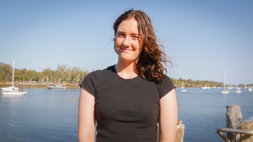 A woman with curly brown hair in the a black tshirt smiling. River, boats and trees in background