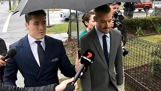 Two men, under an umbrella, walking into court with journalists nearby.