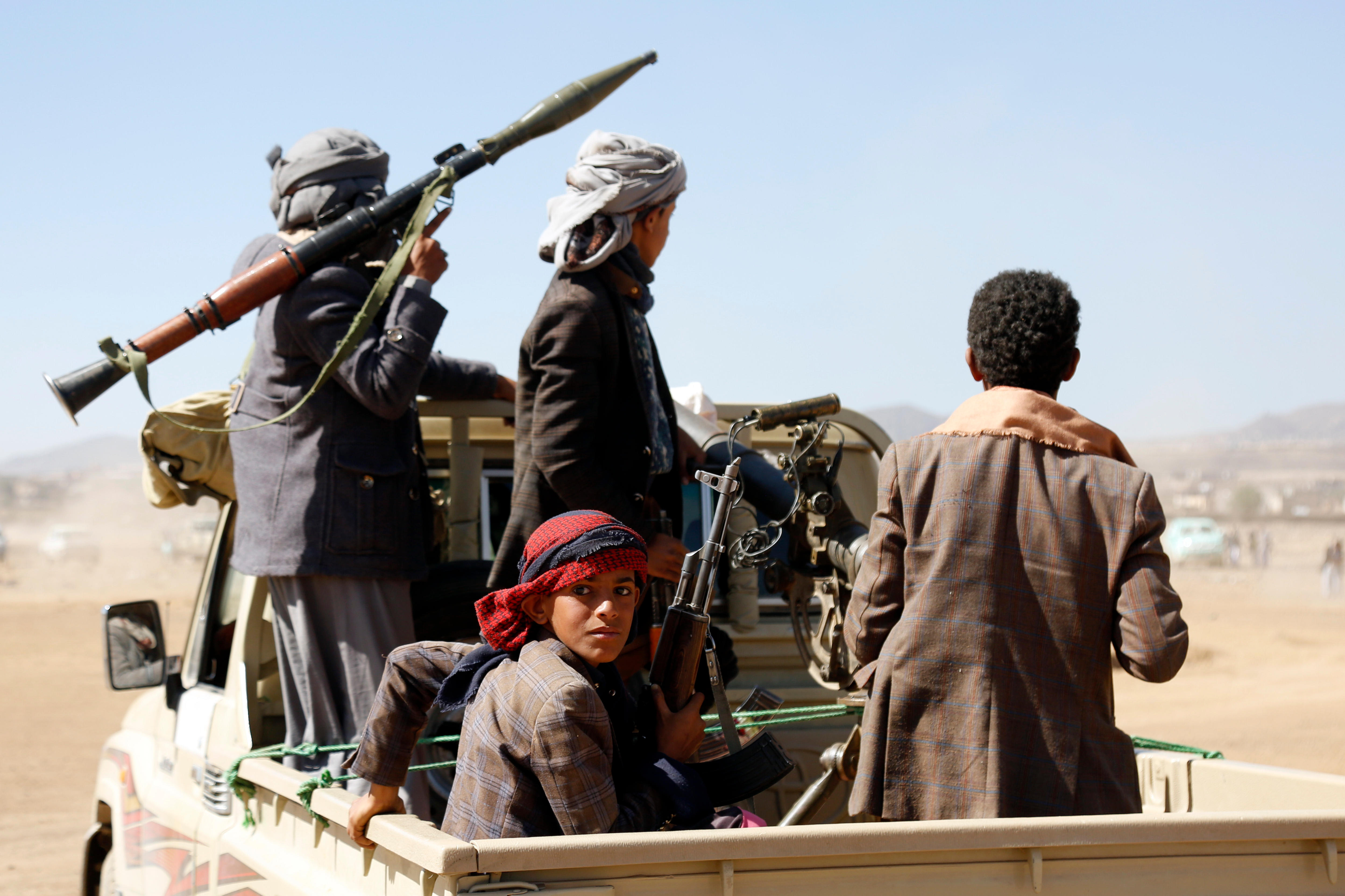 A medium shot of four men sitting or standing in the back of a ute in the desert. Some carry guns, one carries rocket launcher.