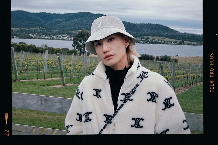 A person wearing a white jacket and hat is standing in front of vines and a river.