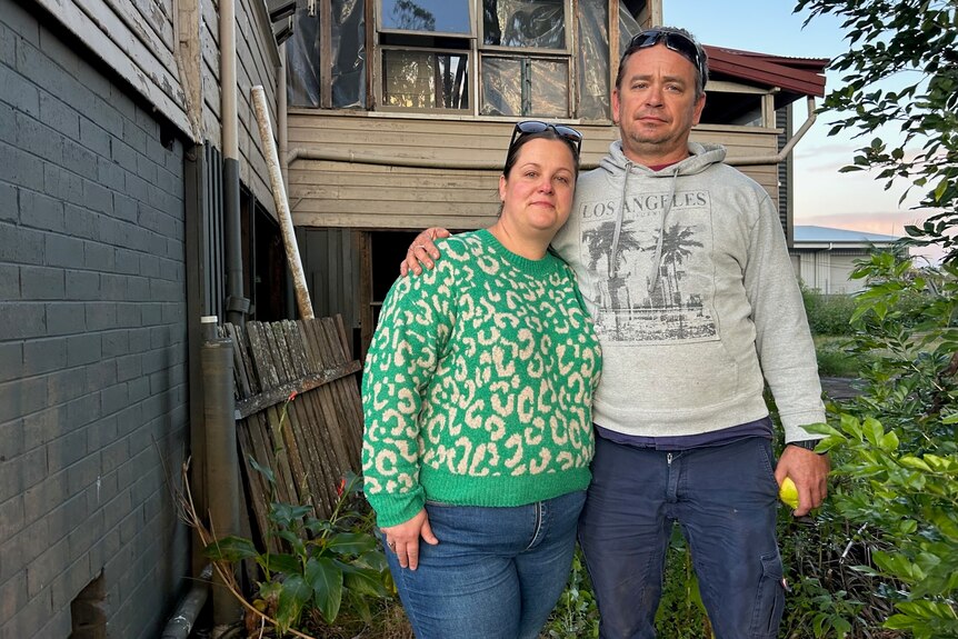 couple wearing jeans and jumpers outside a damaged timber home 