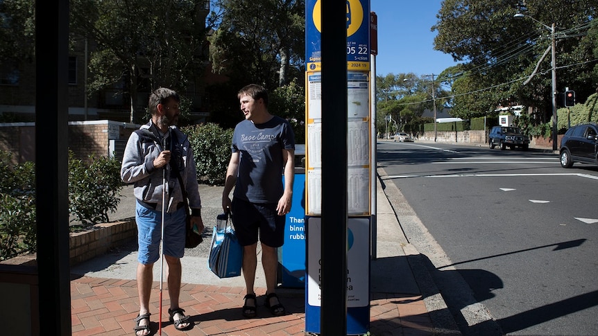 Two men stand and talk at a bus stop in a suburban street. One man holds a white cane.