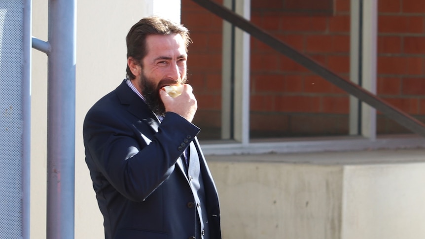 Bearded man wearing suit eats apple in front of retro court building.