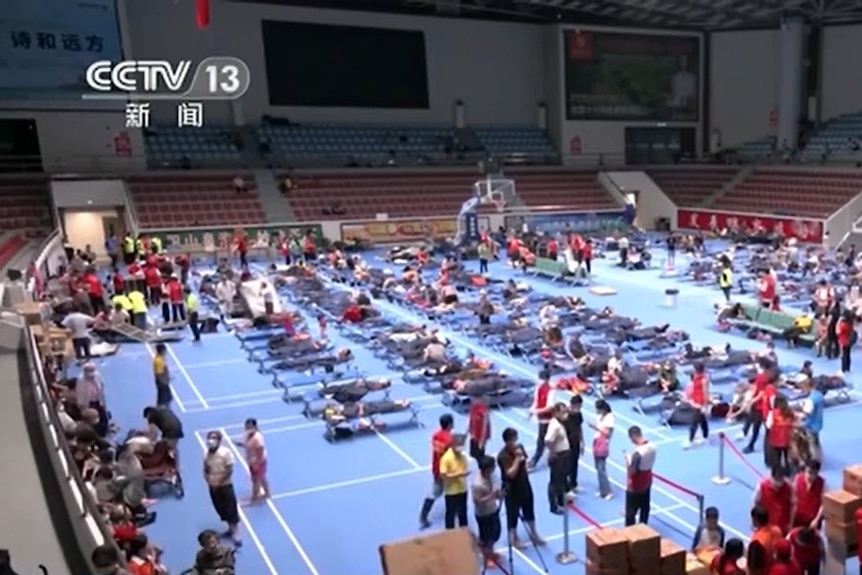 A wide shot of an indoor sports complex, now full of camping beds. People mill around the room.