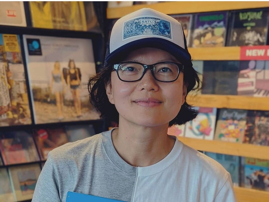 A woman with glasses and a hat, holding a vinyl record, smiling at the camera.