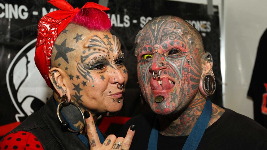 A woman and a man with facial tattoos, piercings and eyeballs tattooed black and yellow