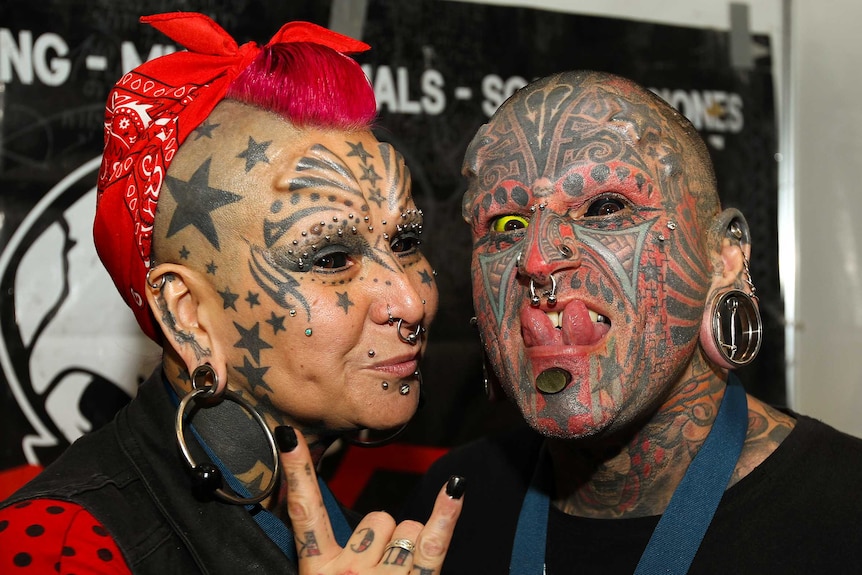 A woman and a man with facial tattoos, piercings and eyeballs tattooed black and yellow