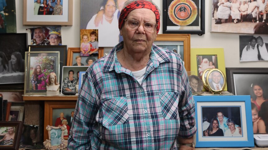 An elderly woman in a red headscarf stands in front of a wall of family photos