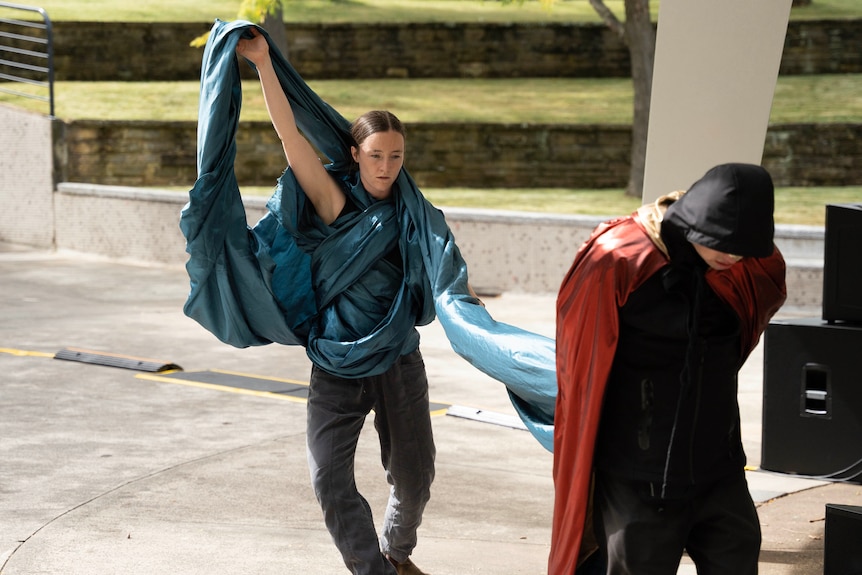 Woman with a green tentacle made from material wrapped around her follows a man in a hood 