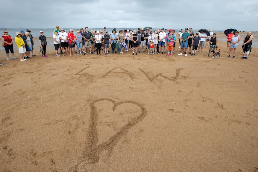 The word 'law' written in the sand with about 40 people standing behind it in a rally following the murder of Emma Lovell.