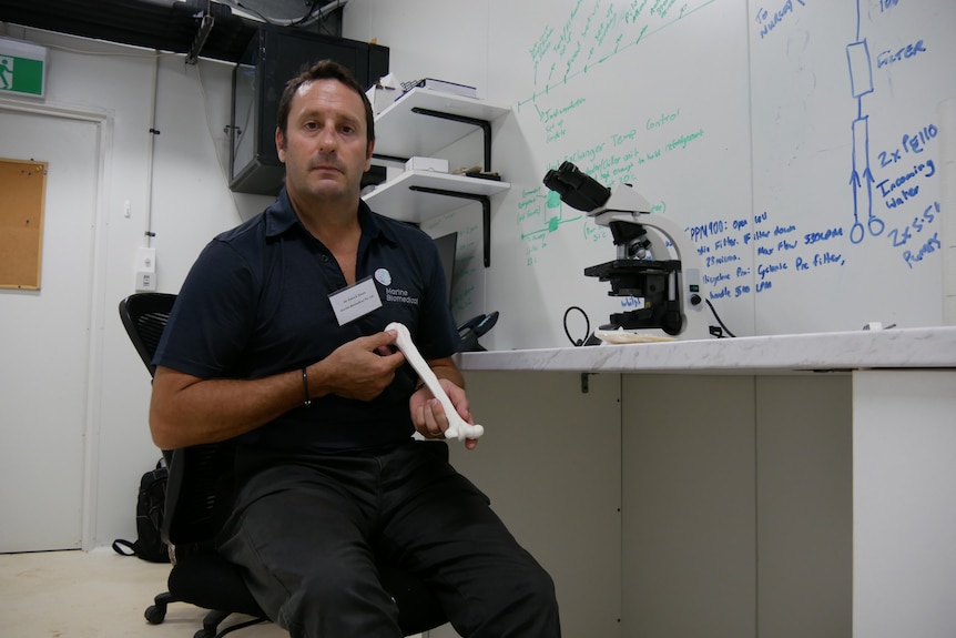 A man sits in front of a microscope and holds a bone in front of him