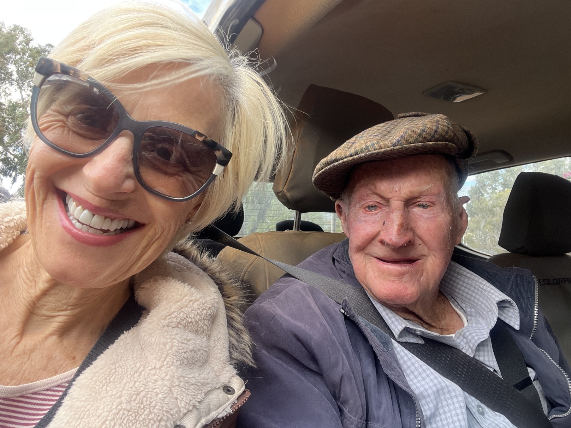 a blonde short haired lady with sunglasses smiling with an old man with a hat on smiling at the camera