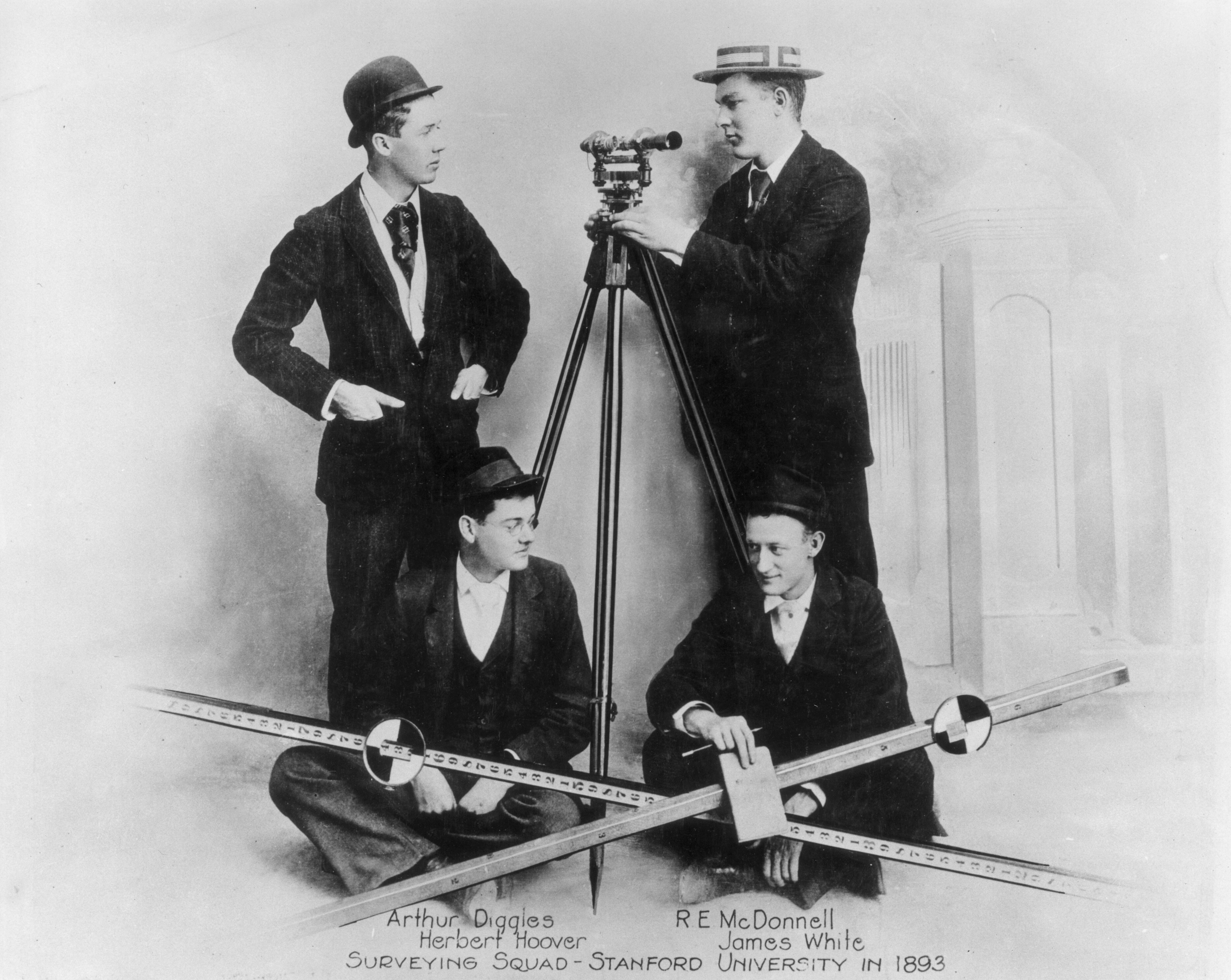 A black and white photo of four well-dressed students with surveying tools.
