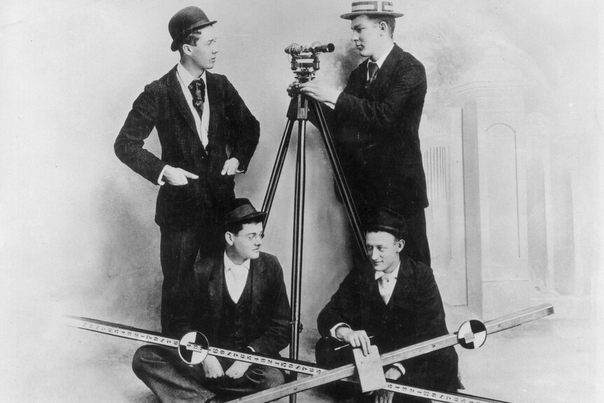 A black and white photo of four well-dressed students with surveying tools.
