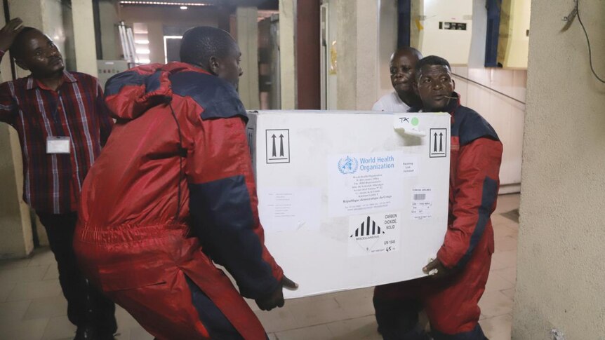 Two men in red carry large white box containing vaccines