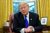 Donald Trump sits at the President's desk in the Oval Office, his hands clasped in front of him, glancing down towards the desk.