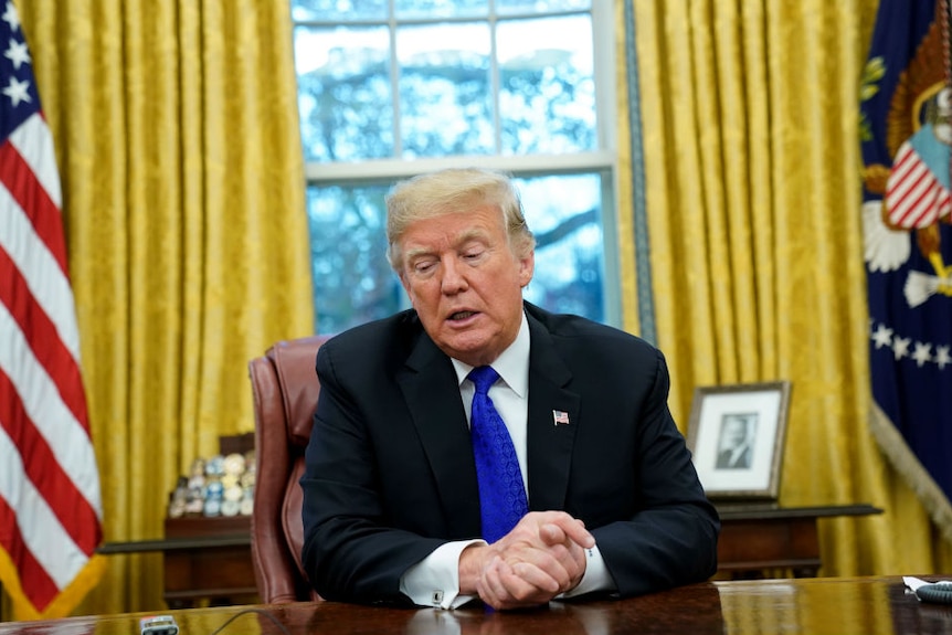 Donald Trump sits at the President's desk in the Oval Office, his hands clasped in front of him, glancing down towards the desk.