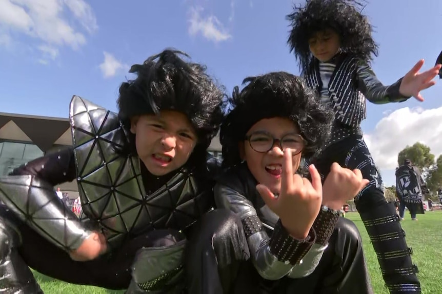 Three boys wear black rock and roll star style wigs and KISS rock band costumes and pull rock star poses close to the camera.