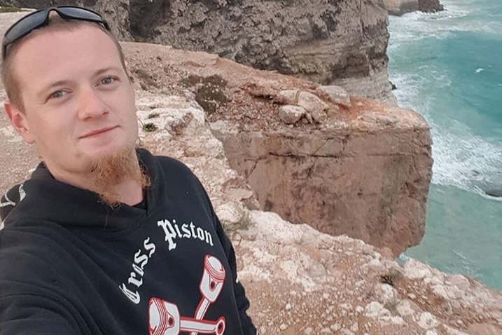 Luke Dempster stands near a cliff taking a selfie photo wearing a black jumper with water seen below the cliff.