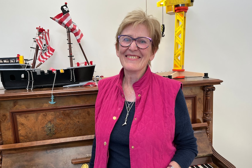 An older, bespectacled woman stands in front of a piano, smiling warmly.